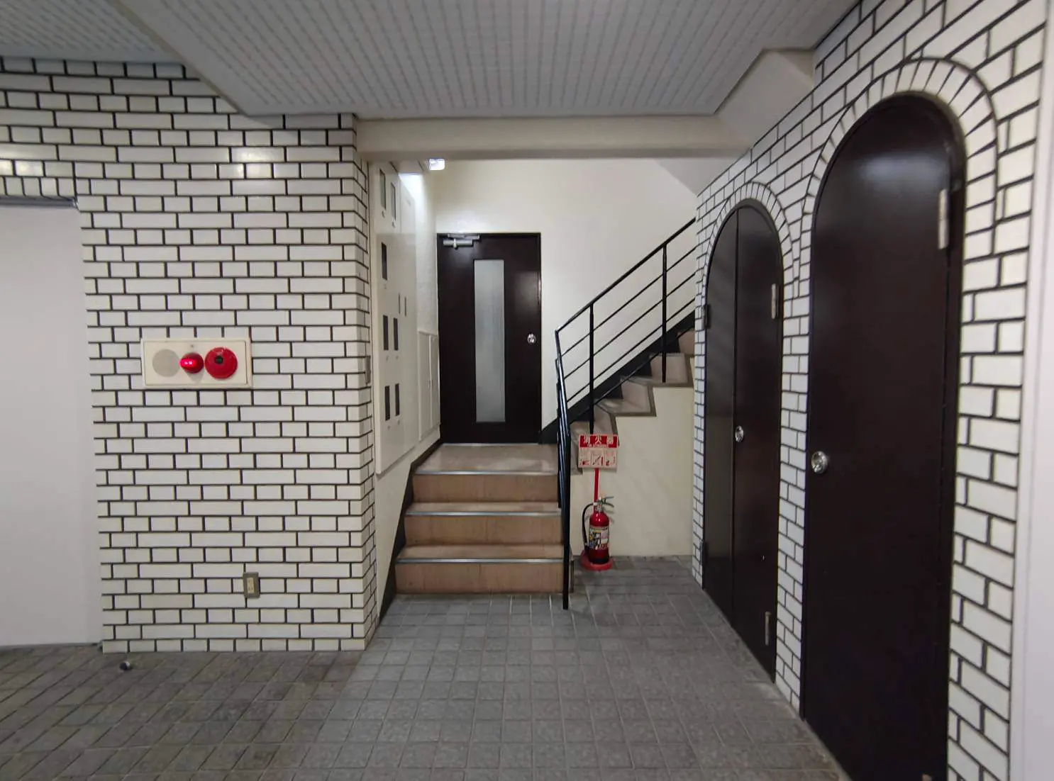 - レンガ調のタイルで覆われたエントランスホール、木製扉と階段、消火器が設置された安全対策のある空間