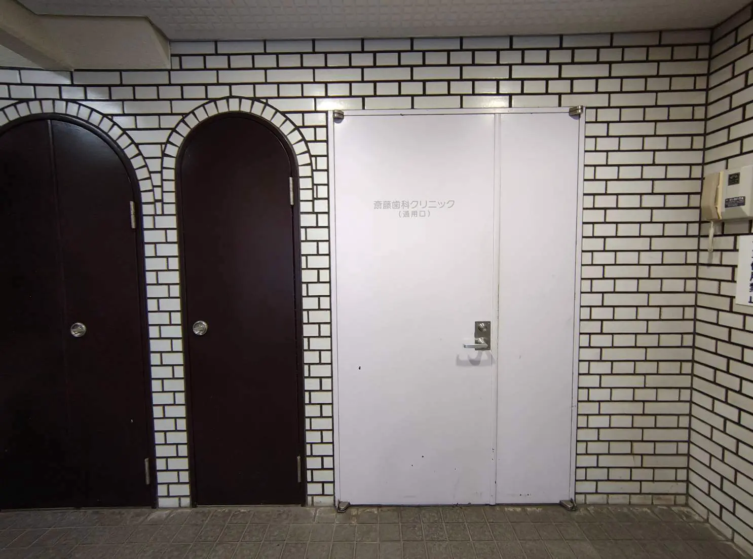 - 煉瓦模様の壁に囲まれた歯科クリニックの入口、白い扉と左右に配置された暗色の扉が特徴の空間