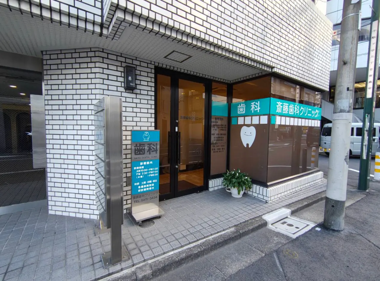 - 斉藤歯科クリニックの外観、白いタイル張りの建物と入口、看板に描かれた歯のイラストが特徴の歯科医院