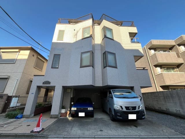 - 白いタイル張りの外観を持つ3階建てのアパートメント、屋上テラス、2台の車が駐車されている駐車スペース、晴れた青空の背景
