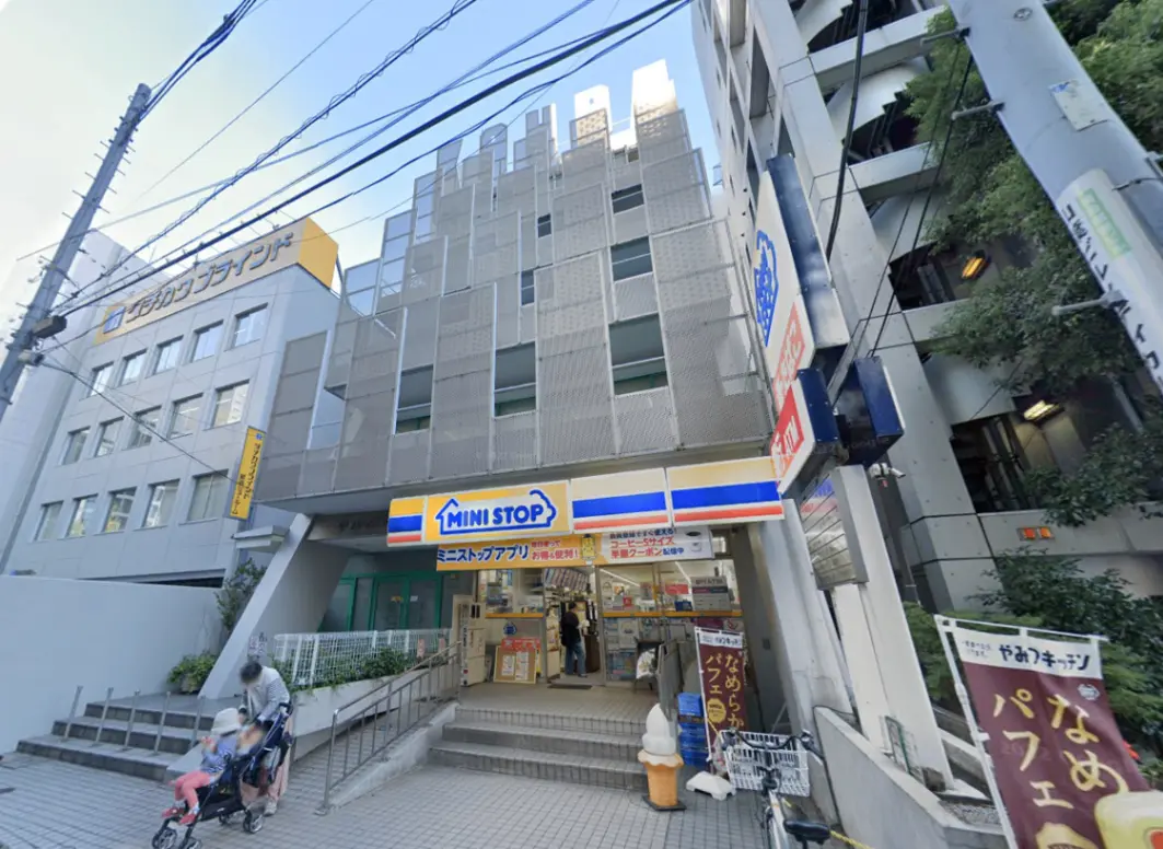 - ミニストップの店舗外観、階段を上る親子連れと周囲の高層ビル、明るい設計と看板の様子