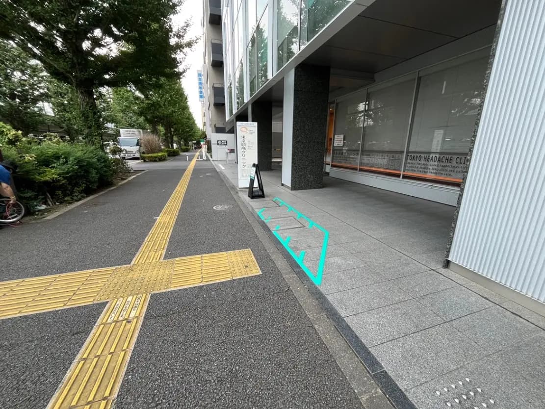 - 都市の街路に面したクリニックのエントランス、青いラインが引かれた歩道と点字ブロックがある風景
