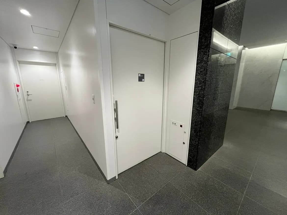 - 白い壁とグレーのタイルの廊下、エレベーターの扉とセキュリティシステムがあるビルの入口部分