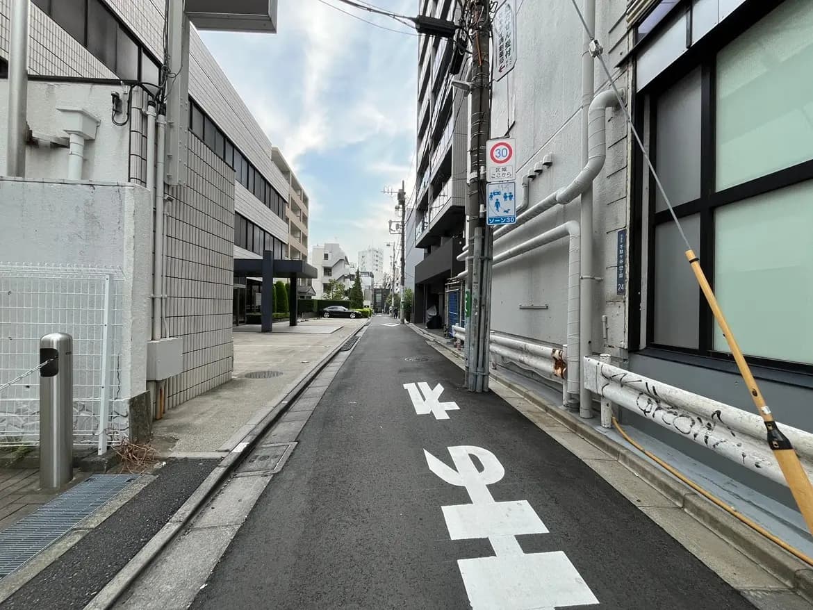 - 都市の裏道、両側にビルが並び、白い矢印の交通標識とガードレールが見える静かな風景