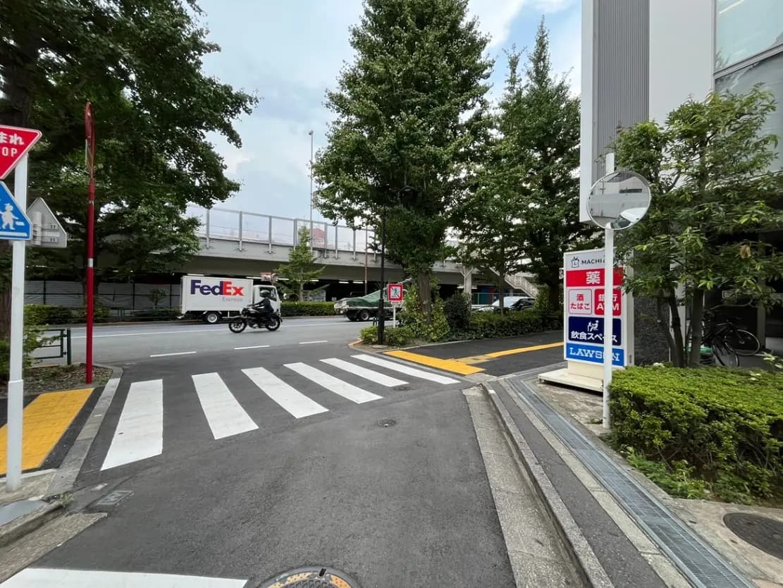 - 駐車場の通りに面した交差点、FedExのトラックが走り、横断歩道がある都市の風景、周囲には緑の樹木と店舗の看板が見える