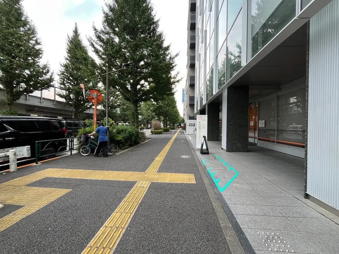 - 自転車を押している男性と沿道の緑の木々、舗装された歩道、周囲に立ち並ぶ現代的な建物の風景