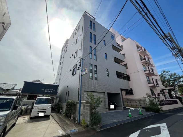 - モダンなアパートの外観、白いタイル張りの壁と青空、隣にはクリーニング店の看板が見える通りの風景