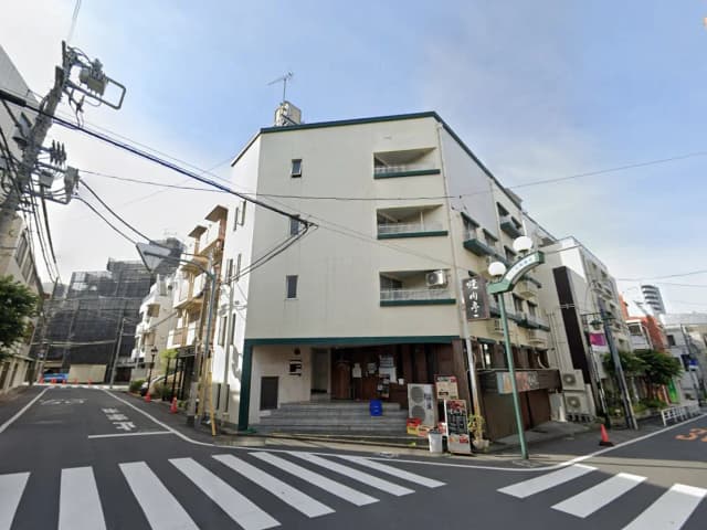 - 街角に位置する外観が白い居酒屋、階段の前に看板が掛かり、周囲に電柱と電線が見える景観