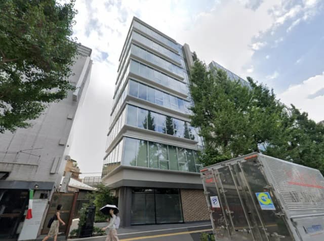 - 近代的なガラス張りのオフィスビル、周囲に緑が豊かで、通行人と貨物車が写る街角の風景