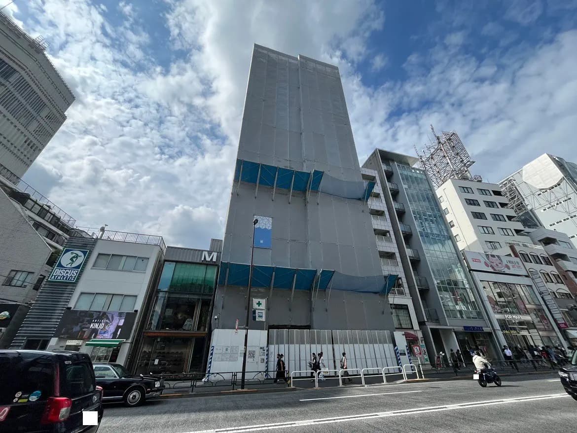 - ビルの改装中の外観、工事用シートがかかった高層ビルと周囲の店舗、青空と雲が広がる風景