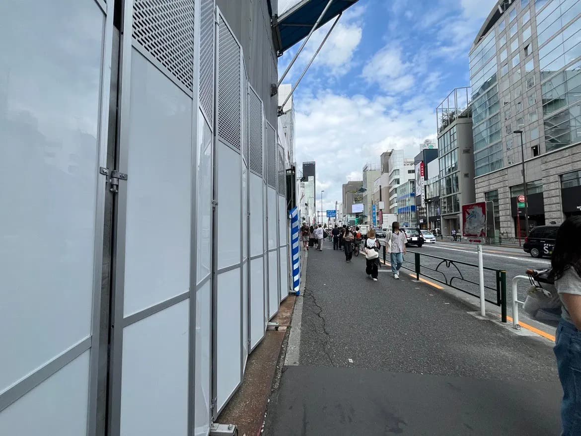 - 都市の通り沿いに立つ白い仮設壁、混雑した歩道を行き交う人々と美しい青空が広がる風景