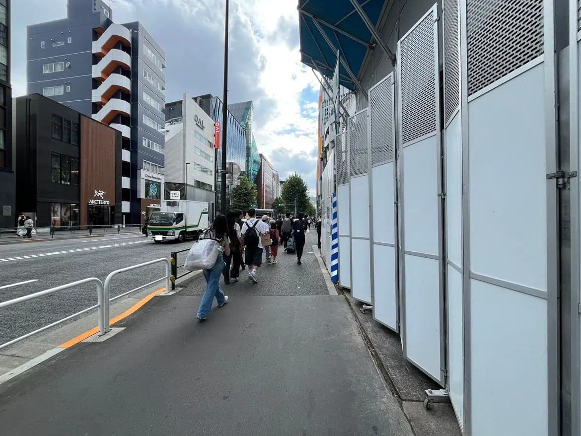 - 都市の歩道を歩く人々、背景には店舗が並び、建物の外壁に白い仮設のフェンスが立てられた風景