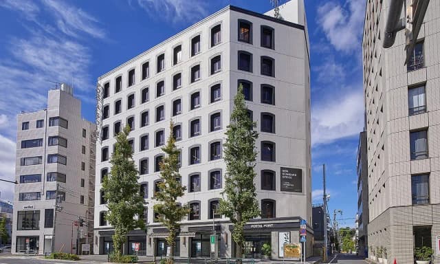 - 現代的なデザインのオフィスビル、周囲に高い木々と他の建物が並ぶ街の風景