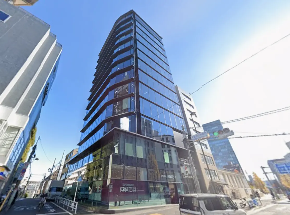 - 現代的なデザインのガラス張りビル、周囲には商業施設や道路があり、青空を背景にした都市景観