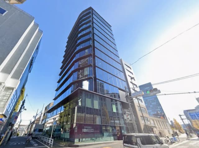 - 現代的なデザインのガラス張りビル、周囲には商業施設や道路があり、青空を背景にした都市景観