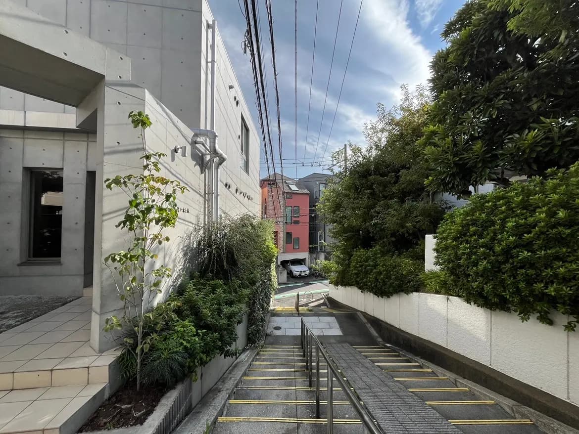 - コンクリートの外壁を持つ現代的な建物の側面、緑豊かな植栽、遠くに見える街並みと青空の風景