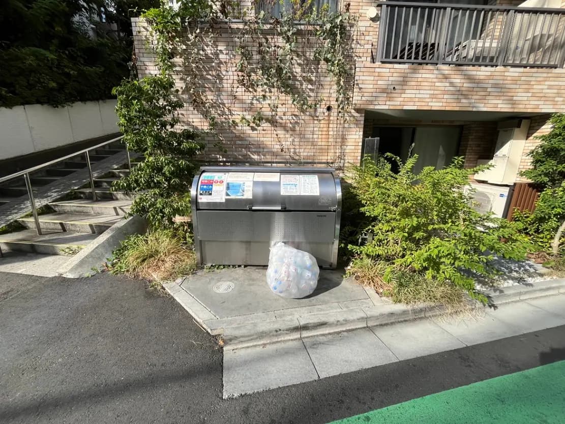 - マンション前に設置された清掃用ゴミステーション、周囲に緑の植栽と市道の風景が広がる様子