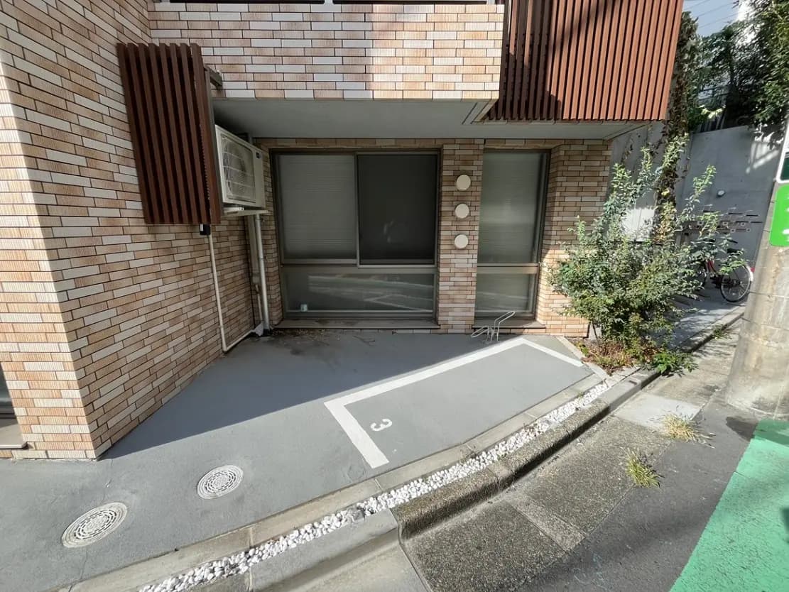 - モダンな外観の建物、レンガ調の壁と大きな窓、駐車スペースには番号が付けられている様子、周囲には緑の植栽が見える