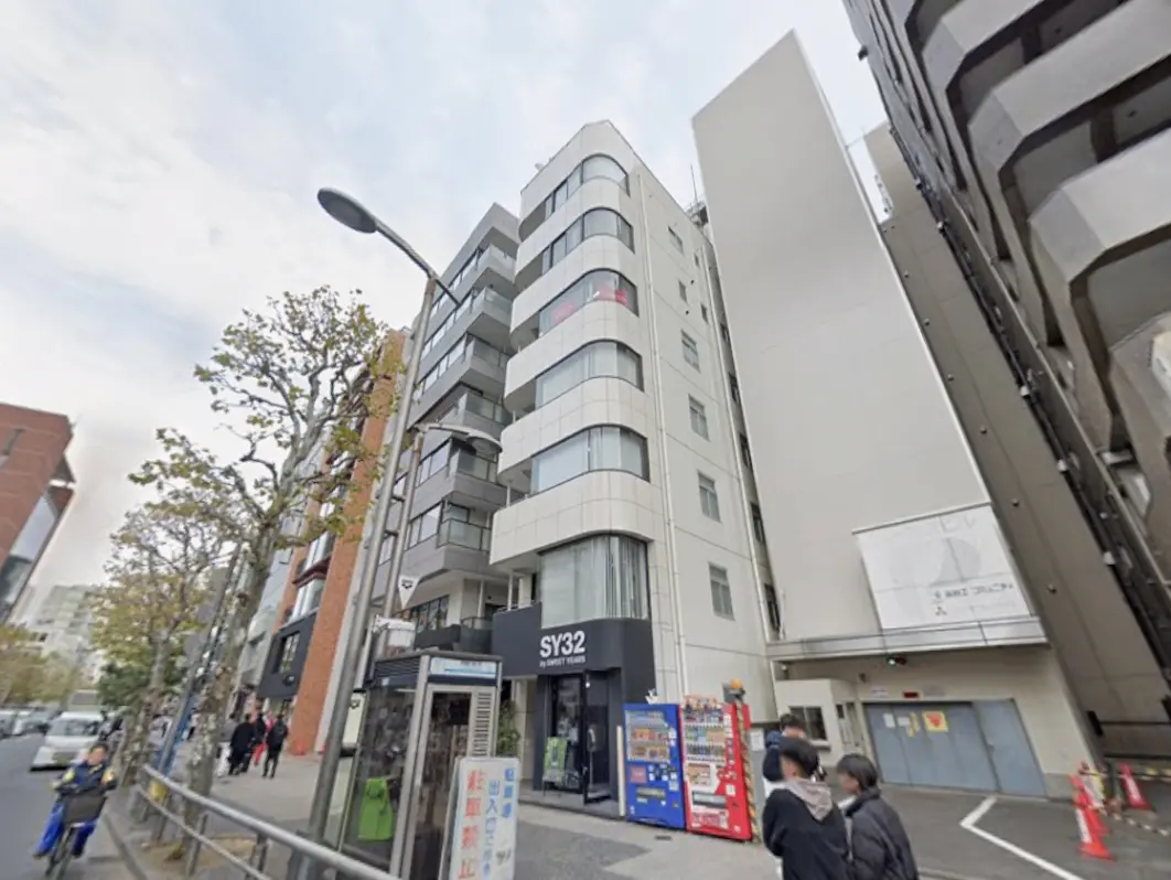 - 都会の住宅ビル、シンプルな外観、道路沿いに人々が行き交う風景、サイドに自転車を止めた人がいる画像