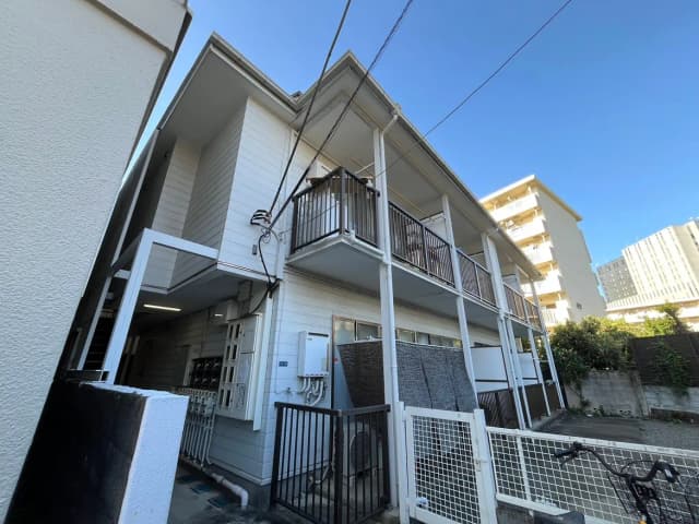 - 明るい空の下にある、木造の2階建てアパートの外観、バルコニーが特徴で周囲の建物とともに写っている全景