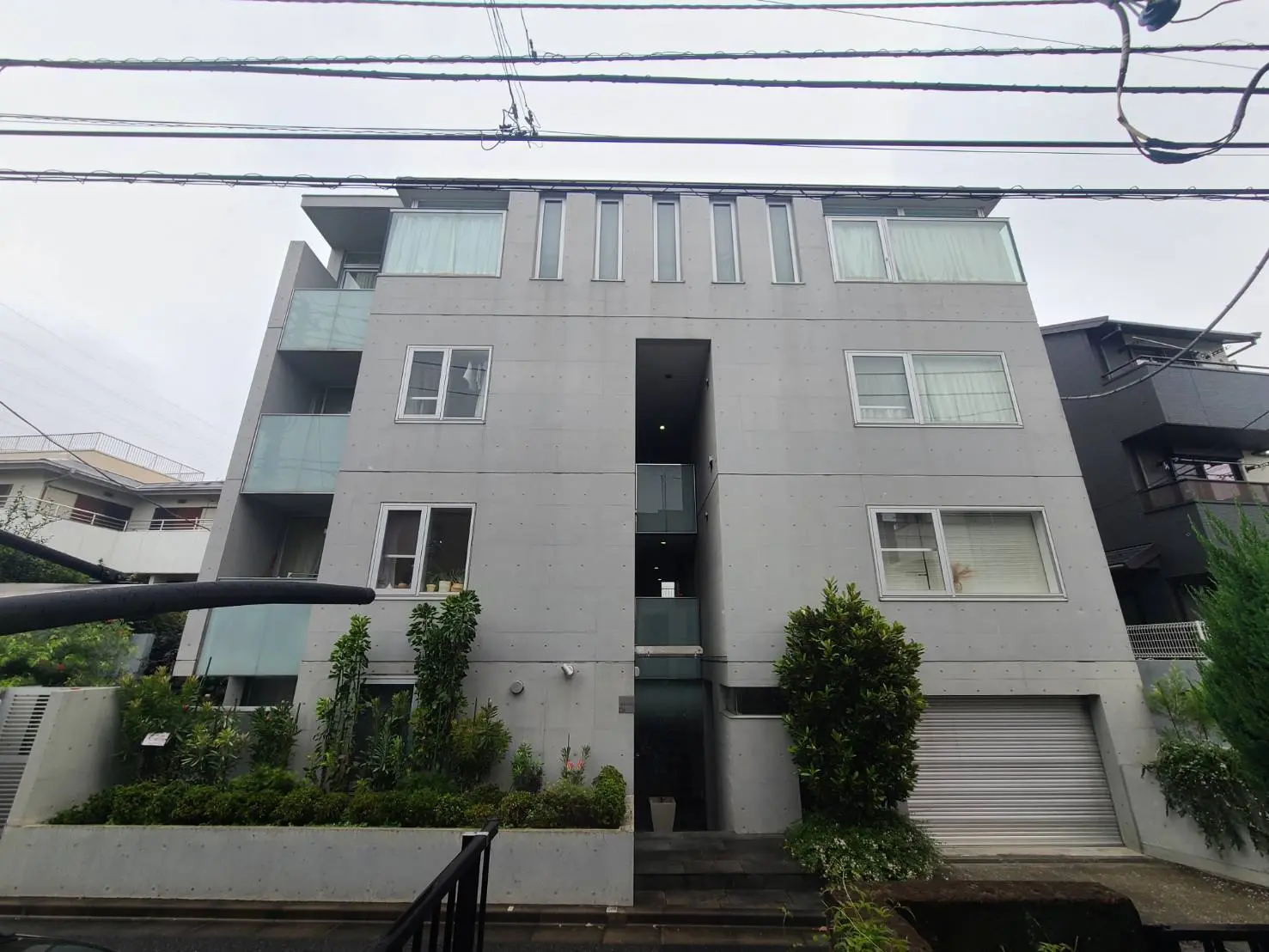 - モダンなコンクリート製のマンション外観、ガラスのバルコニーと緑豊かな植栽が特徴的な住宅ビル