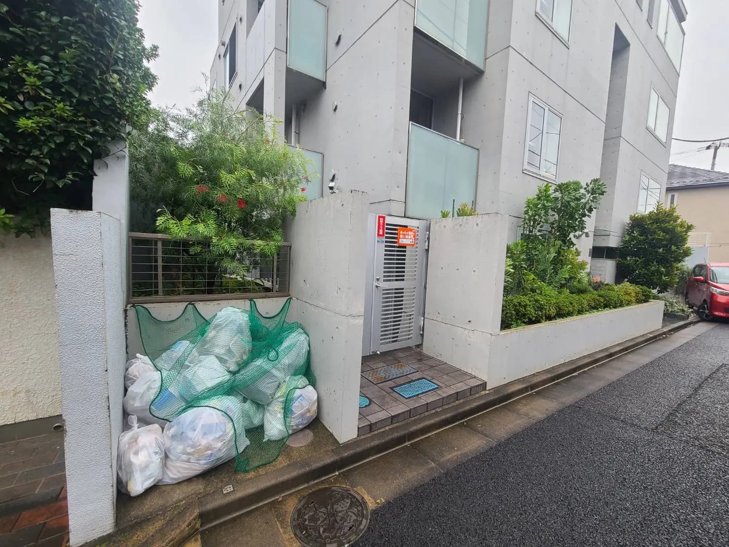 - コンクリート造の集合住宅とその前に置かれたゴミ袋、緑豊かな植物とエントランスの様子