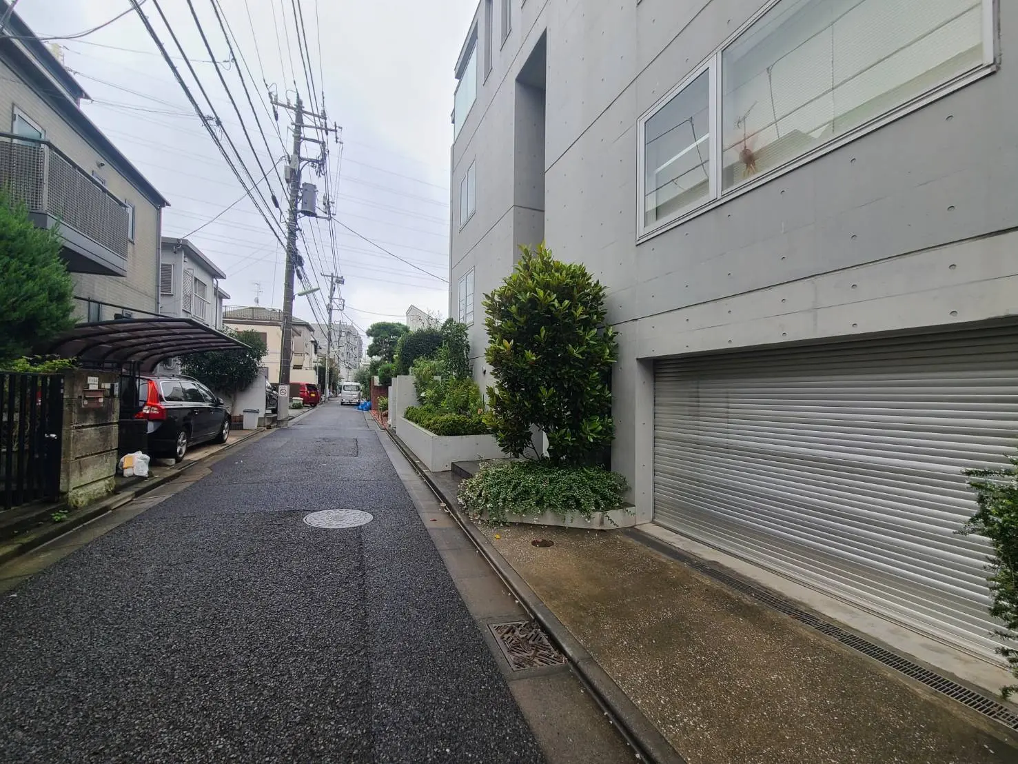 - コンクリート製の現代的な建物と狭い道路、周囲に豊かな緑や車が停車している静かな住宅街の風景