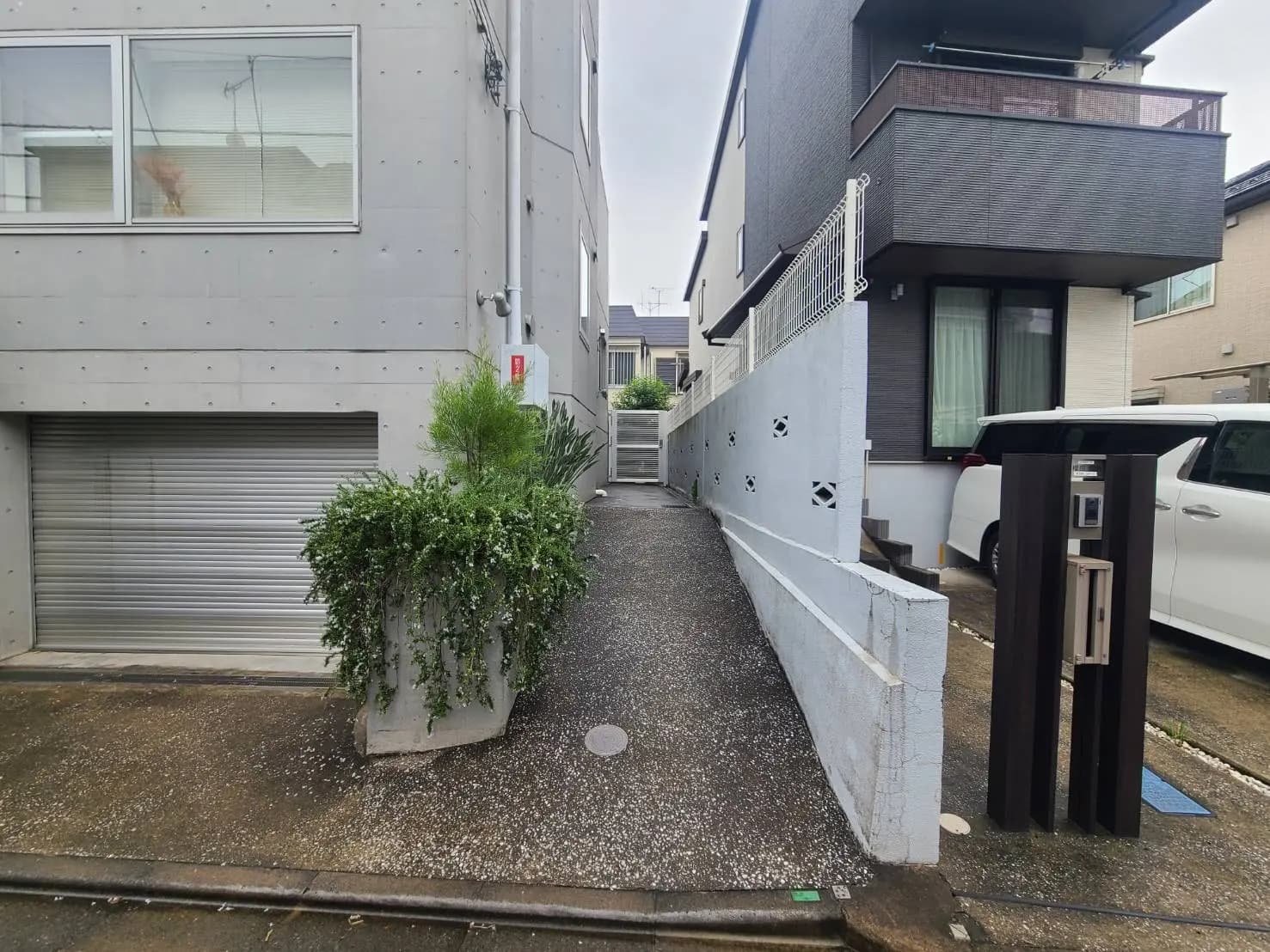 - コンクリートの壁に囲まれた住宅の側道、シンプルなデザインの植栽とシャッター付きガレージ、白い塀と自動車のある風景