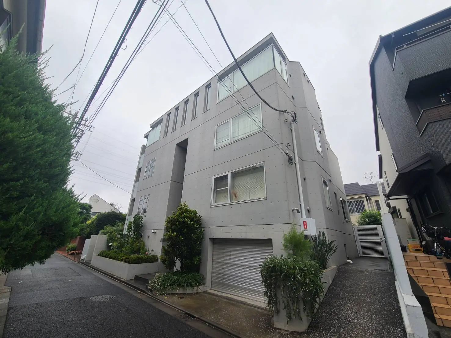 - コンクリート外壁のモダンな建物、周囲に植栽があり、隣の建物と接近した住宅街の風景