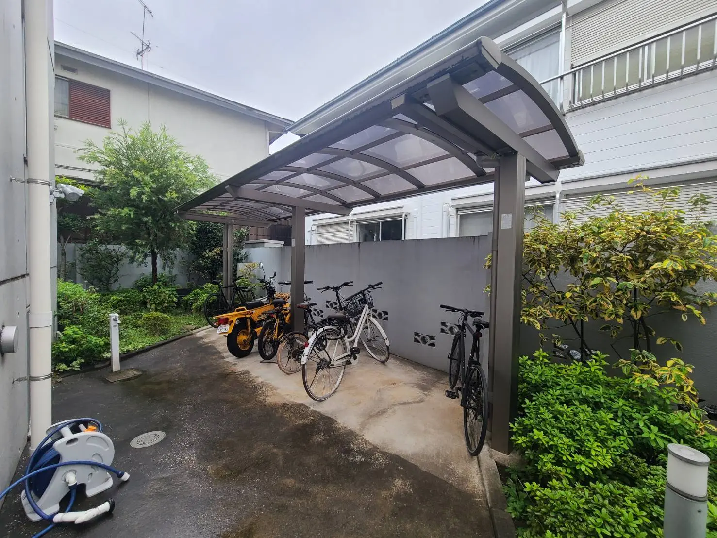 - 自転車とバイクが駐輪されているカバー付きの駐輪場、周囲には緑豊かな植物が育つ庭がある風景