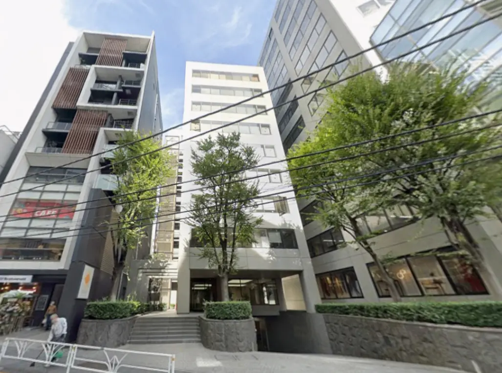 - 現代的なデザインの複合ビル、緑豊かな木々に囲まれたエントランス、周囲にはカフェや店舗が立ち並ぶ都市風景