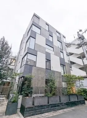 - モダンなデザインの外観を持つ建物、白とグレーのタイル張り、周囲には緑の植栽が施された街中の様子