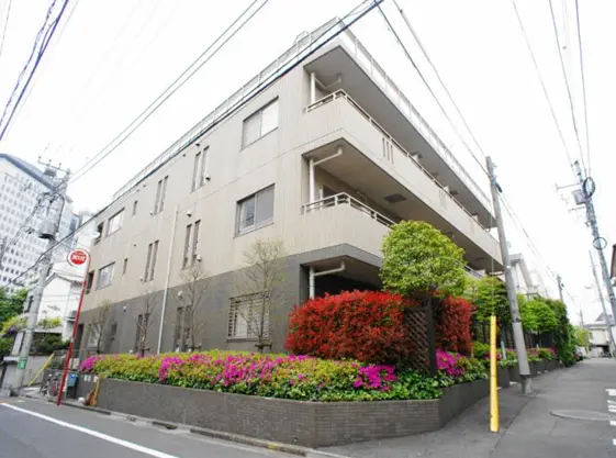 - 角度から撮影されたマンションの外観、色とりどりの花壇と緑の植栽がある道路沿いの風景