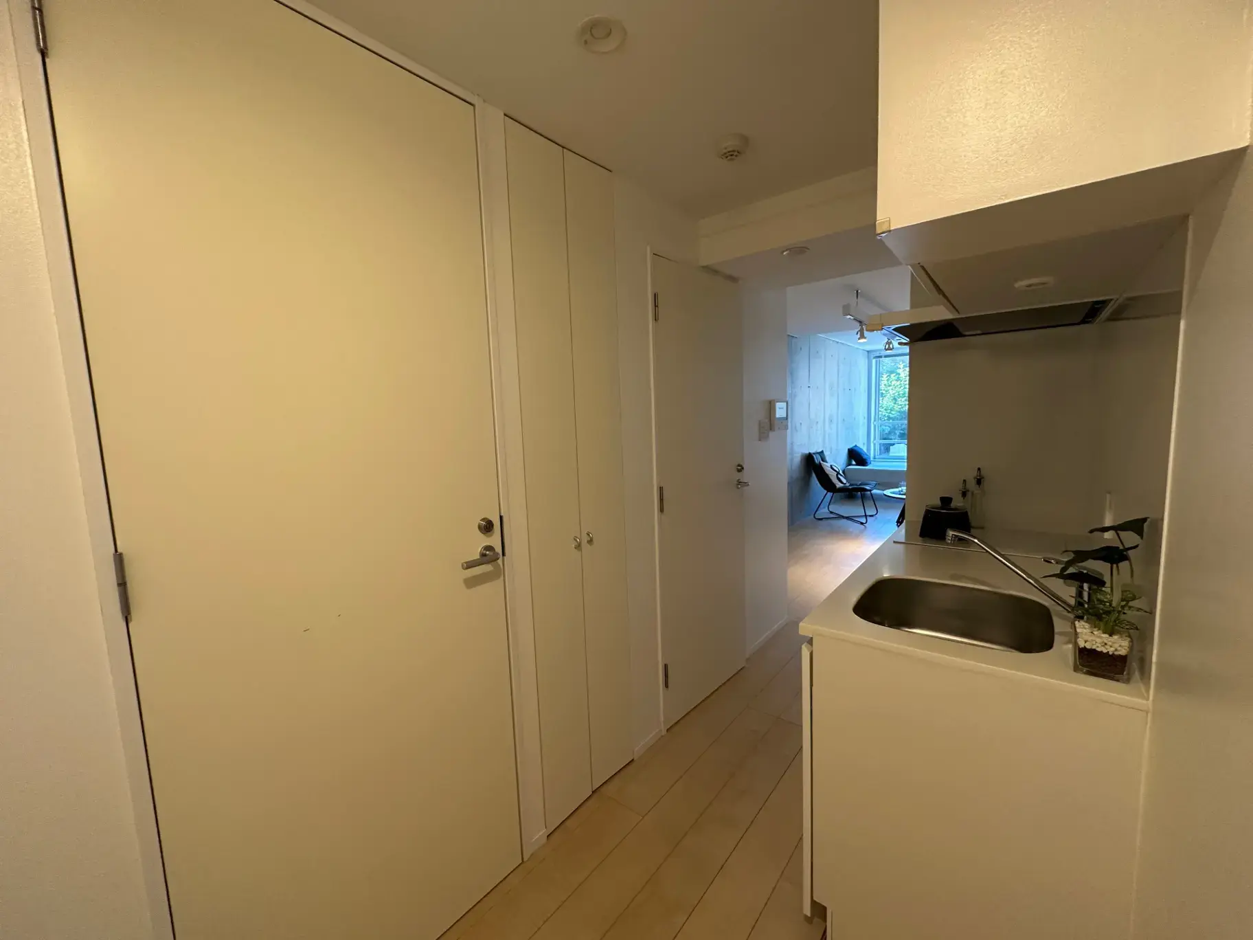 シンプルなデザインのキッチンと白い扉が特徴の明るい廊下、奥に見えるリビングにはモダンな椅子が配置された室内写真