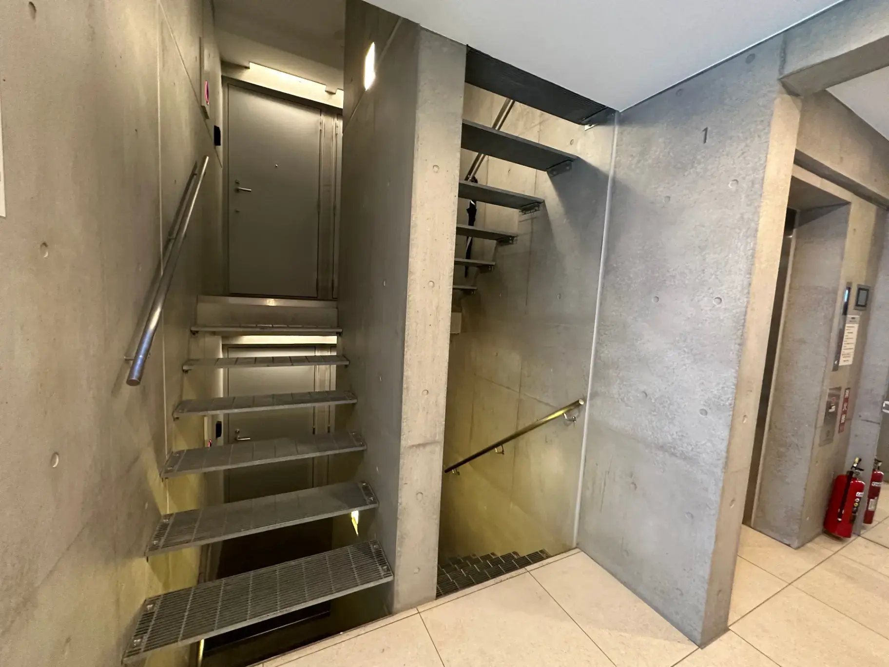 コンクリートの壁と金属製の手すりを備えたモダンなデザインの階段、照明と消火器が設置された非常階段の入り口