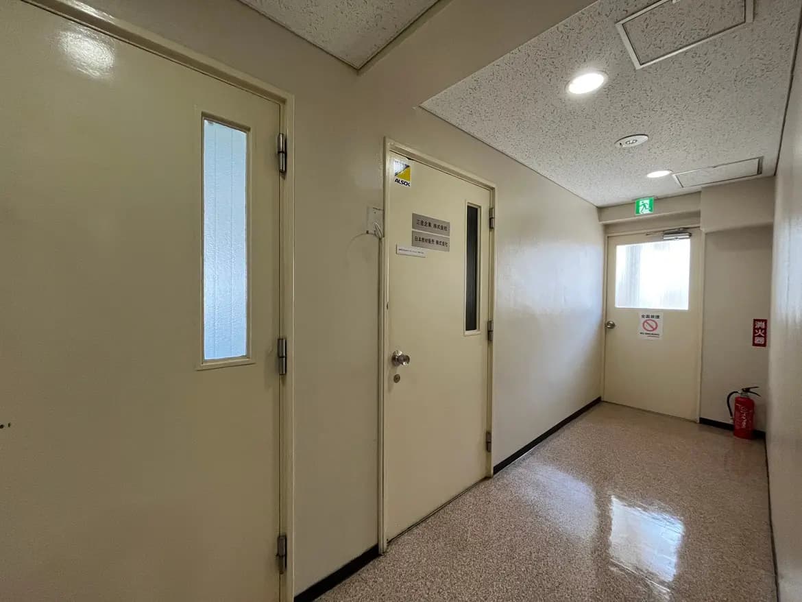 - 明るい色調の廊下、左側に二つのドア、右側に窓のある出口、非常口と消火器の表示があるインテリア