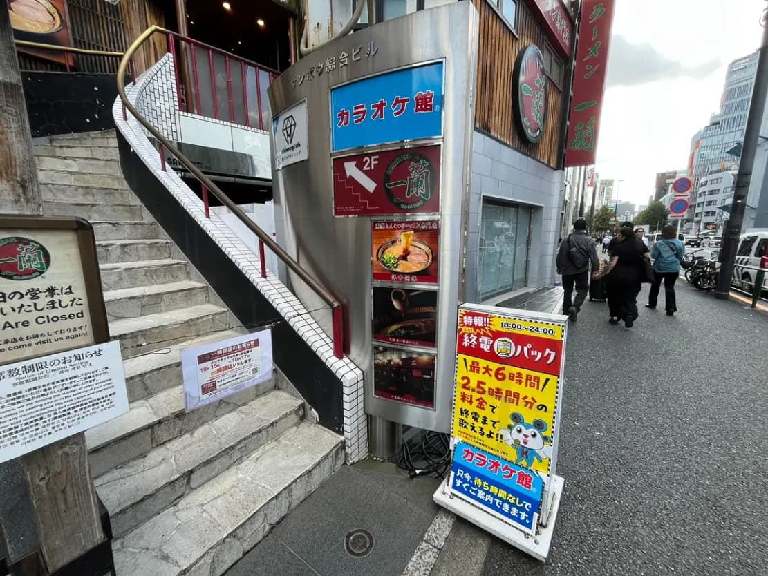 - 繁華街に位置するカラオケ館の入口、階段を上がる際の外観、看板が目立つビルの周辺の様子