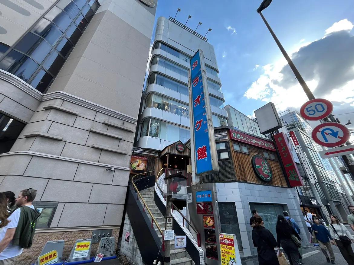 - カラオケ館の外観、近くにラーメン店の看板、繁華街の様子と人々が行き交うショッピングエリア