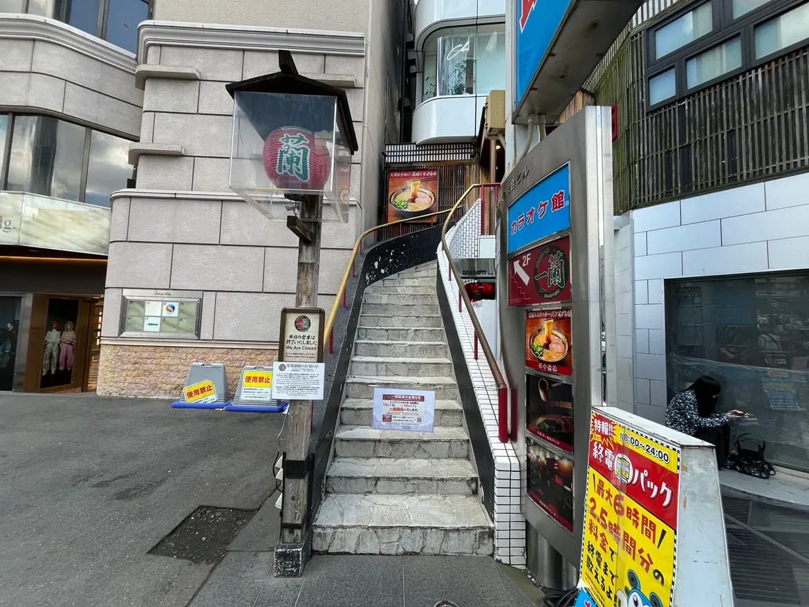- 商業ビルの入り口、階段を上ると見えるラーメン店の看板とカラオケの案内、周辺に掲示された注意書きの立て看板