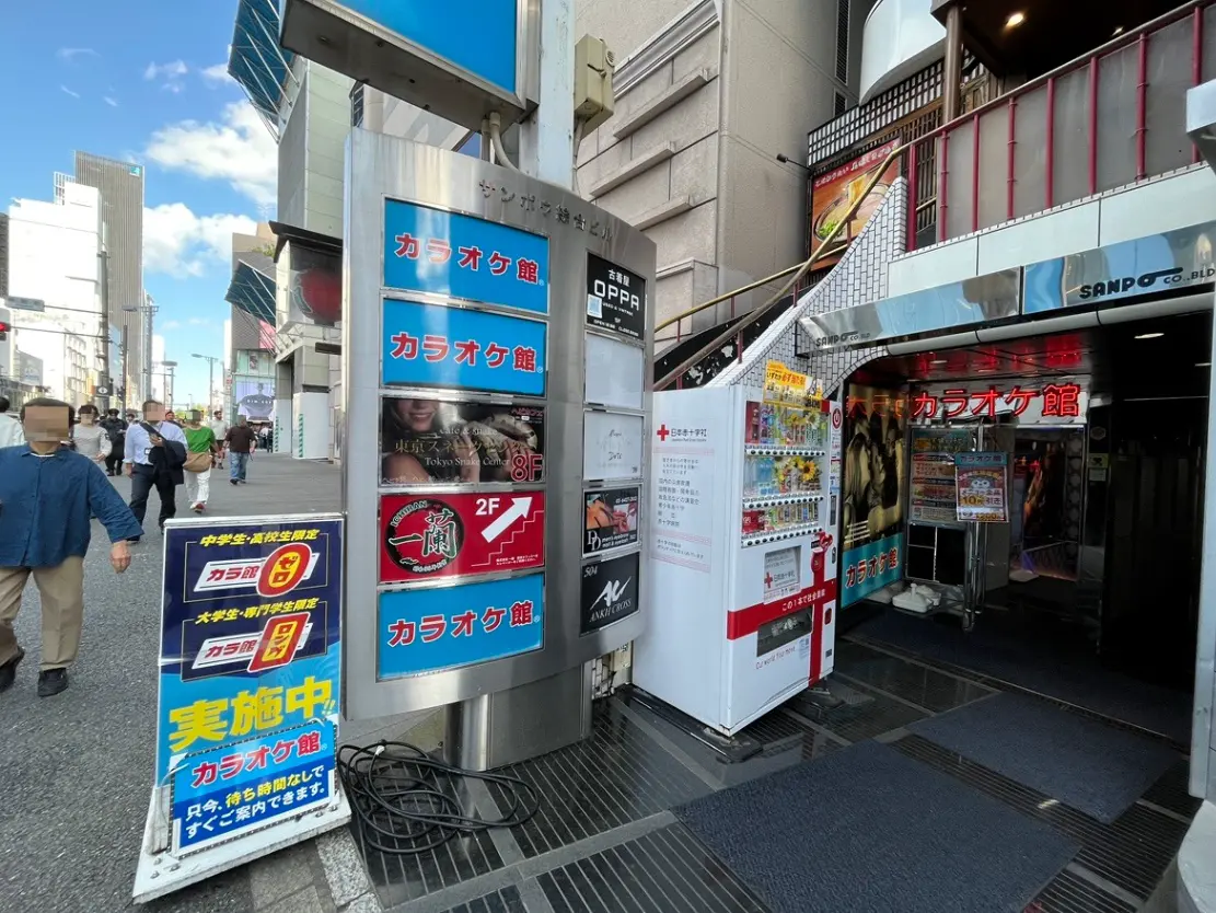 - カラオケ館の入り口、周囲には人々が行き交う賑やかな通り、ガイドサインやポスターが並ぶ商業エリアの風景
