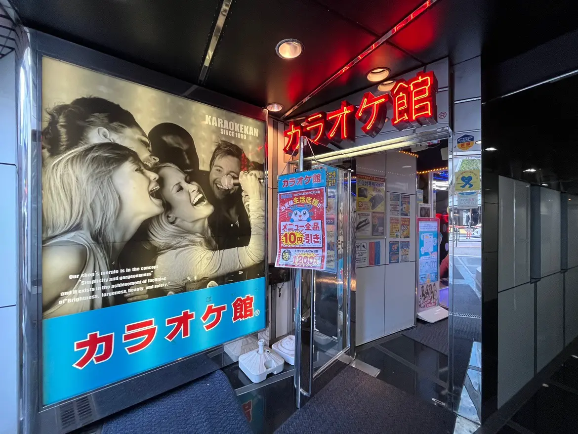 カラオケ館の入口、ネオン看板が輝く外観と併せて宣伝ポスターが掲示された様子、活気あるカラオケ文化を感じさせるシーン