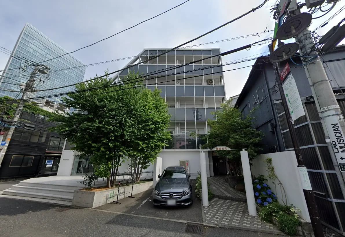 現代的なビルの外観、白い外壁に大きな窓、前に緑豊かな木、黒い車が駐車している都市風景