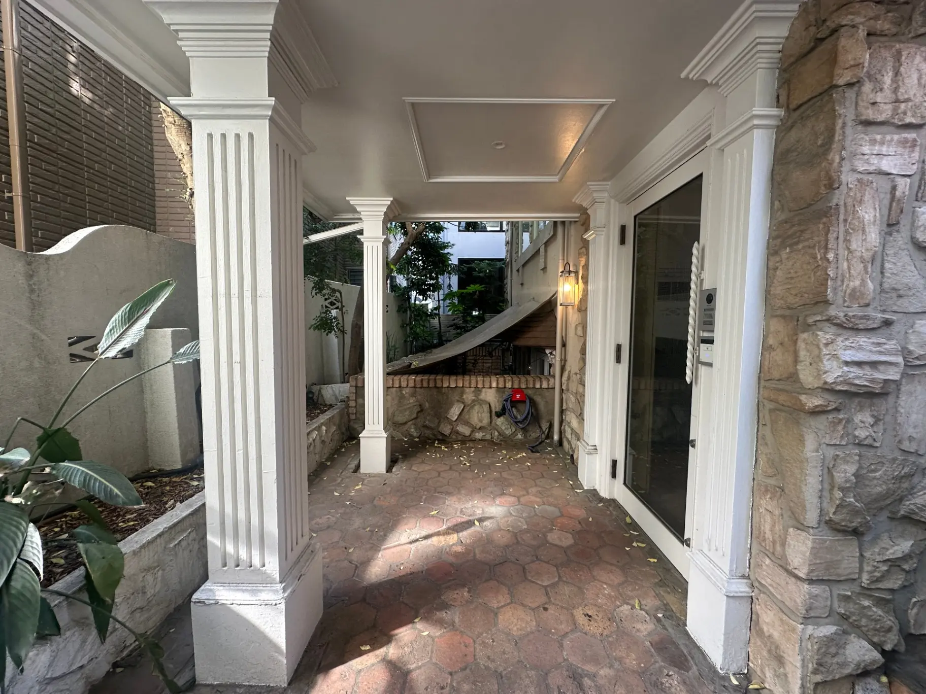 白い柱と石造りの壁が特徴的なエントランス、レンガ敷きのアプローチと植物が彩る住宅の玄関