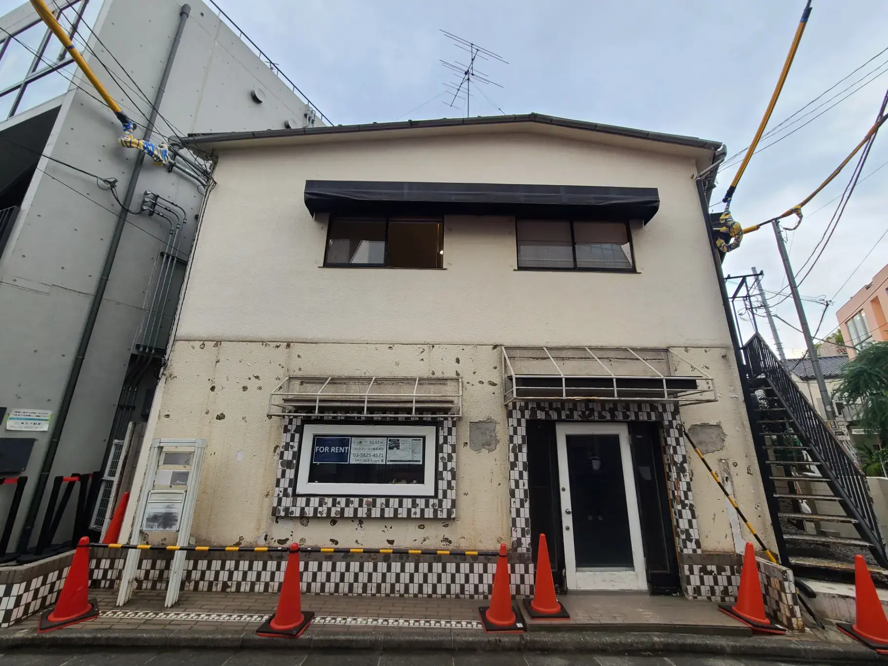 "賃貸物件の募集看板がある古い二階建ての建物の外観、工事用の交通規制コーンが前に設置されている"
