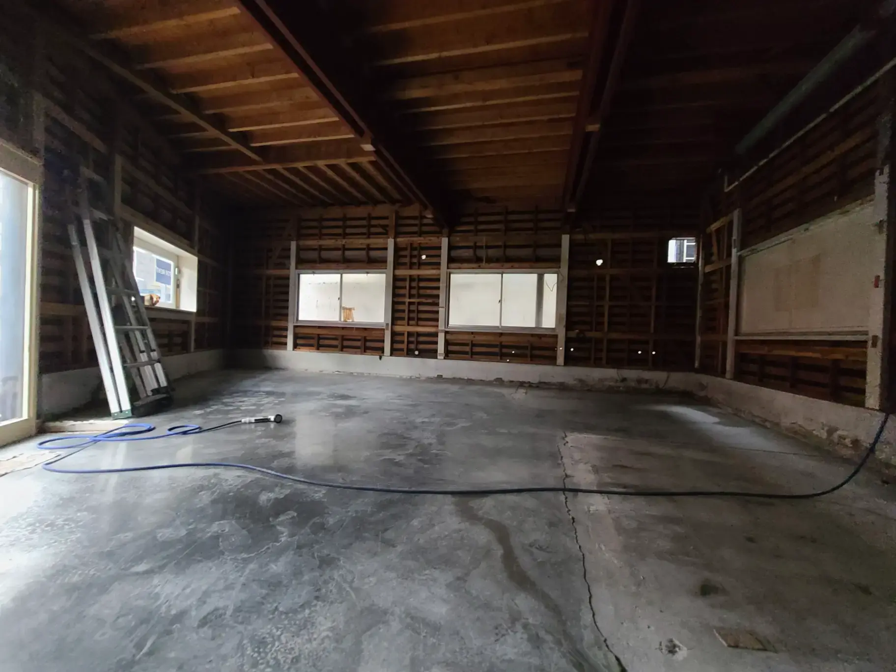 「改装前の空の倉庫内部、コンクリートの床にはホースが敷かれ、背景にははしごが立てかけられている」