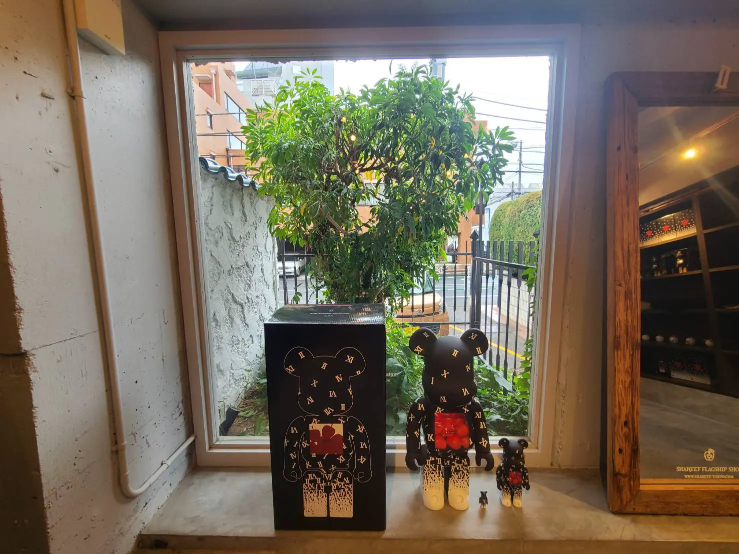 ウィンドウディスプレイに展示されたデザイナーズアートフィギュア、木製の枠とガラスの窓、背景には緑豊かな植物