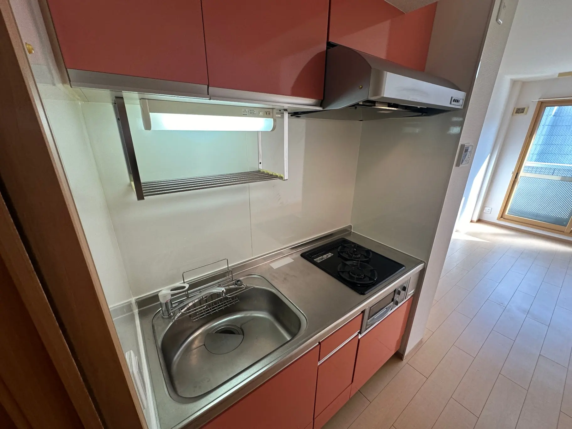 コンパクトなキッチン、ステンレス製シンクとガスコンロ、レッドカラーキャビネット、白い壁と木製フローリングのインテリア