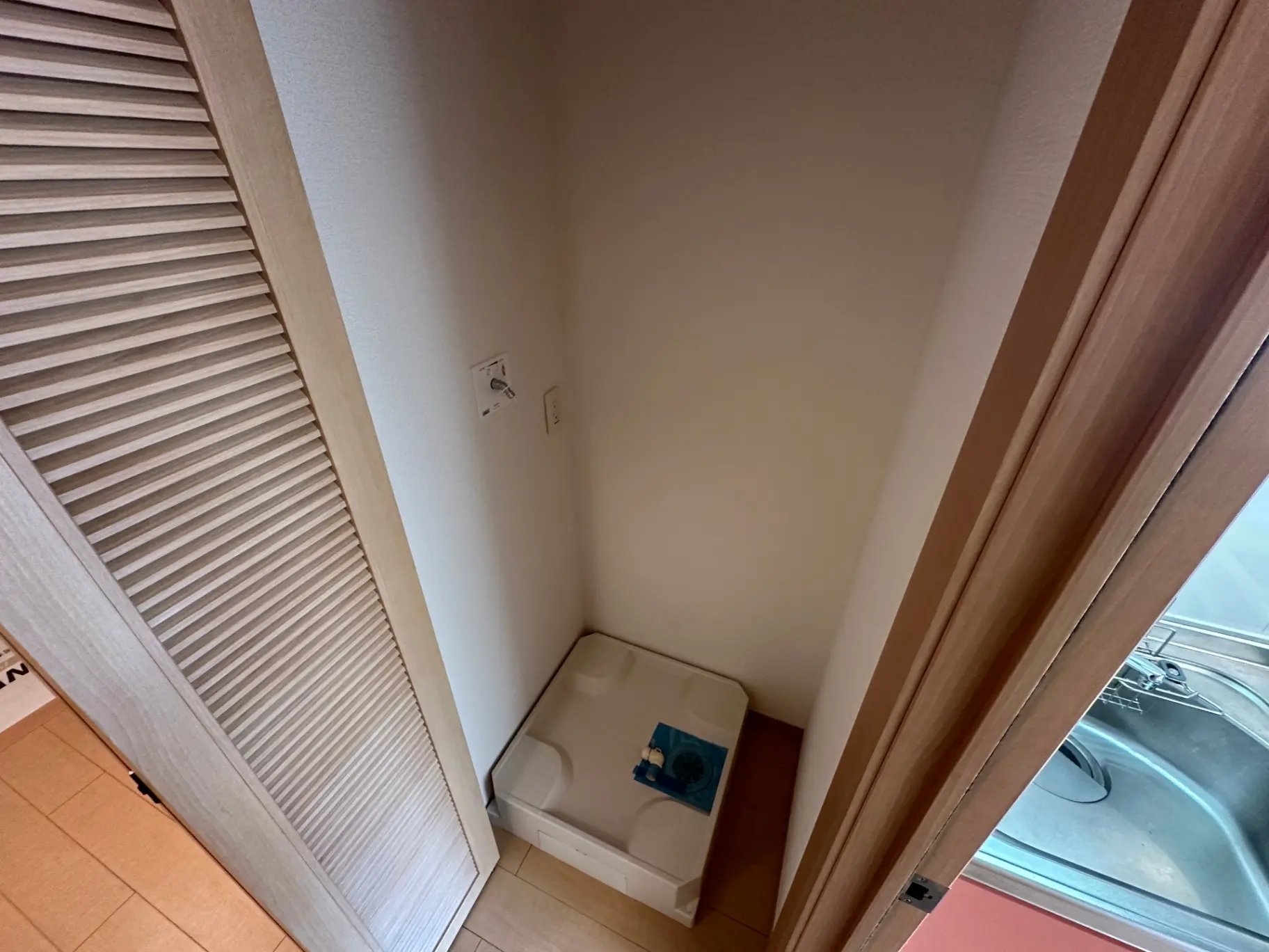 狭い洗面所の角に置かれた白い洗濯機、隣にはスライド式のクローゼット扉、タイルの床とシンクの一部が見える家庭用設備