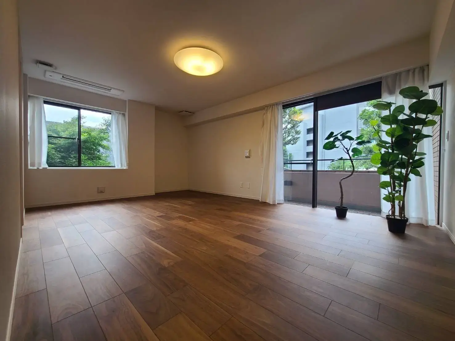 明るい空間のリビングルーム、木目調のフローリング、白いカーテンの窓とバルコニーへの出入口