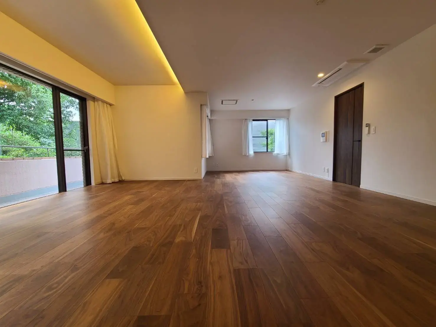 広々としたリビングルーム、ダークブラウンの木製フローリング、自然光が差し込む大きな掃き出し窓と白い壁のインテリア